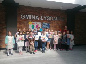 Dzieci biorące udział w konkursie pozują do zdjęcia na tle ściany z cegieł, na której widnieje napis GMINA ŁYSOMICE a pod nim przyczepiona grafika dwojga dzieci z plecakami na plecach.