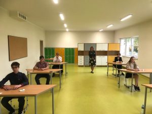 Uczniowie siedzą w ławkach w oczekiwaniu na egzamin. Ławki ustawione są w dwóch rzędach. Po środku stoi nauczycielka będąca członkiem komisji nadzorującej egzamin.