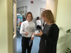Uczeń wraz z nauczycielką będącą przewodniczącą komisji egzaminacyjnej stoją w wejściu do sali. Uczeń trzyma w dłoni kartkę z numerem stolika, a nauczycielka przezroczysty pojemnik z kolejnymi numerami.
