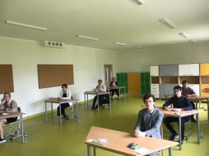 Uczniowie siedzą w ławkach w oczekiwaniu na egzamin. Ławki ustawione są w dwóch rzędach.