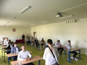 Uczniowie siedzą w ławkach w oczekiwaniu na egzamin. Ławki ustawione są w dwóch rzędach. Przed uczniami stoi nauczycielka będąca członkiem komisji nadzorującej egzamin.