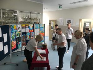 Uczeń wraz z przewodnicząca swojej komisji nadzorującej stoi przy stoliku zakrytym czerwonym obrusem. Za stołem stoi Pani Dyrektor, która komisyjnie rozpakowuje arkusze egzaminacyjne.