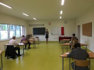 Uczniowie siedzą w ławkach w oczekiwaniu na egzamin. Ławki ustawione są w dwóch rzędach. Po środku stoi nauczycielka będąca członkiem komisji nadzorującej egzamin.