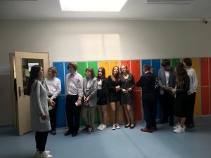 Uczniowie ustawieni w rzędzie pod kolorowymi szkolnymi szafkami, czekają na wejście do sali egzaminacyjnej. Przed nimi stoi nauczycielka będąca przewodniczącą komisji egzaminacyjnej.