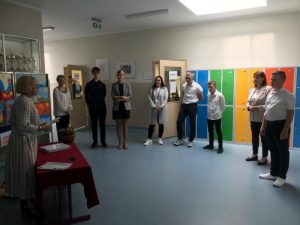 Przedstawiciele ósmoklasistów wraz z przewodniczącymi poszczególnych komisji egzaminacyjnej stoją w półkolu na szkolnym holu. Przed nimi za stołem przykrytym czerwonym obrusem stoi Pani Dyrektor trzymająca w dłoniach arkusze egzaminacyjne.