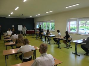 Uczniowie siedzą w ławkach w oczekiwaniu na egzamin. Ławki ustawione są w dwóch rzędach. Po środku stoi nauczycielka będąca członkiem komisji nadzorującej egzamin.