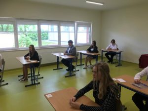Uczniowie siedzą w ławkach w oczekiwaniu na egzamin. Ławki ustawione są w dwóch rzędach.