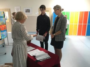 Uczeń wraz z przewodnicząca swojej komisji nadzorującej stoi przy stoliku zakrytym czerwonym obrusem. Za stołem stoi Pani Dyrektor, która komisyjnie rozpakowuje arkusze egzaminacyjne.