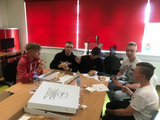 Uczniowie klasy ósmej siedzą przy wspólnym stole i jedzą pizze. Na stole leżą kartony po pizzyi stoja butelki z wodą.