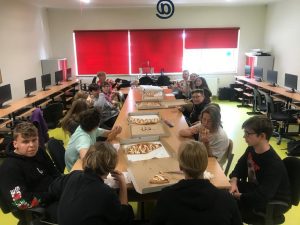 Uczniowie klasy ósmej siedzą przy wspólnym stole i jedzą pizze. Na stole leżą kartony po pizzyi stoja butelki z wodą.