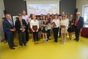 Najlepsi absolwenci jednej ze szkół podstawowych gminy Łysomice pozują do grupowego zdjęcia z rodzicami, wychowawcą, dyrektorem szkoły oraz przedstawicielami gminy. W tle wyświetlone z rzutnika zdjęcie Szkoły Podstawowej w Ostaszewie.
