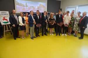 Najlepsi absolwenci jednej ze szkół podstawowych gminy Łysomice pozują do grupowego zdjęcia z rodzicami, wychowawcą, dyrektorem szkoły oraz przedstawicielami gminy. W tle wyświetlone z rzutnika zdjęcie Szkoły Podstawowej w Ostaszewie.
