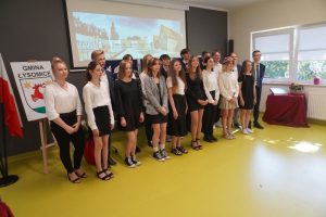 Najlepsi absolwenci ze wszystkich szkół podstawowych w gminie Łysomice pozują do grupowego zdjecia. W tle wyświetla się z rzutnika zdjęcie Szkoły Podstawowej w Ostaszewie.