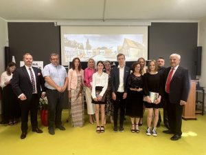 Najlepsi absolwenci Szkoły Podstawowej w Świerczynkach pozują do grupowego zdjęcia z rodzicami, wychowawcą, dyrektorem szkoły oraz przedstawicielami gminy. W tle wyświetlone z rzutnika zdjęcie Szkoły Podstawowej w Ostaszewie.