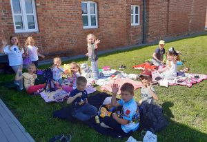 Dzieci z oddziału przedszkolnego piknikują na kocykach rozłożonych na trawniku przed budynkiem szkoły. Uczniowie machają do zdjęcia.
