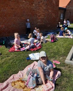 Dzieci z oddziału przedszkolnego piknikują na kocykach rozłożonych na trawniku przed budynkiem szkoły. Na pierwszym planie widać siedzącą na kocyku uczennicę w czapecce z daszkiem na głowie.