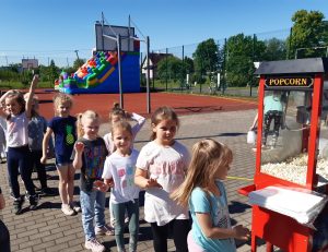Dzieci z oddziału przedszkolnego stoją w kolejce do maszyny do popcornu. W tle widać jedną z ustawionych na szkolnym orilku dmuchanych zjeżdżalni.
