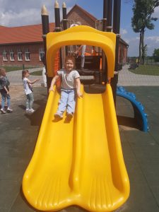 Dziewczynka z oddziału przedszkolnego zjeżdża na żółtej zjeżdżalni ustawionej na szkolnym placu zabaw. W tle widać inne dzieci.