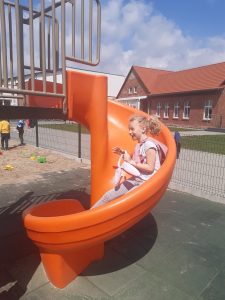Uczennica oddziału przedszkolnego zjeżdża na zawiniętej po łuku, pomarańczowej zjeżdżalni ustawionej na szkolnym placu zabaw.