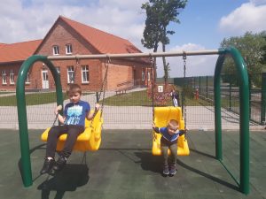 Dwóch uczniów z oddziału przedszkolnego buja się na żółtych huśtawkach ustawionych na szkolnym placu zabaw. W tle widać budynek szkoły.
