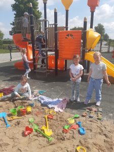 Dzieci z oddziału przedszkolnego na szkolnym placu zabaw - w piaskownicy rozłożono zabawki do zabawy w piasku. Przed piaskownicą stoją dwie uczennice a za ich plecami widać zjeżdżalnię.