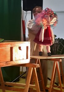 Zdjęcie wykonane podczas występu teatru. Na drewnianym stoliku za którym stoj aktor oparta jest kukiełka świnki w kolorowej peruce.
