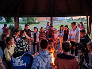 Uczniowie klasy czwartej stoją wokół ogniska rozpalonego pod zadaszoną wiatą stojąco w pobliżu szkoły. Uczniowie trzymają w dłoniach patyki na których umieszczono kiełbaski.