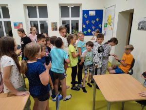 Uczniowie klasy czwartej na szkolnej stołówce stoją w rzędzie i czekają na złożenie życzeń i odebranie cukierka od kolegi, który ma urodziny i siedzi na krzesełku pod ścianą.