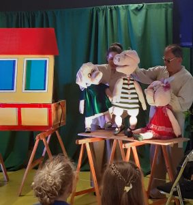 Zdjęcie wykonane podczas przedstawienia teatru. Scenografię stanowią drewniane ławki. Na jednej z nich stoi oparta przednia ściana domku a na drugiej ustawiono trzy lalki świnek ubranych w kolorowe stroje i poruszanych przez dwoje aktorów stojących za nimi.