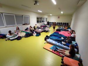 Uczniowie klasy czwartej sziedzą na posłaniach ułożonych pod ścianami sali teatralnej. Uczniowie przygotowują się do snu i są przebrani w piżamy.