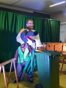 Zdjęcie wykonane podczas przedstawienia teatru. Aktor podrzymuje dużą kukiełkę psa ubranego w kraciaste spodnie i koszulę oraz odblaskową kamizelkę. Drugą dłoń aktor opiera na zielonym kosz na śmieci stojącym przed nim.