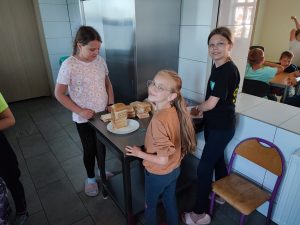 Trzy uczennice klasy czwartej podczas przygotowywania tostów na śniadanie dla całej klasy. Dziewczynki odwracają głowę w stronę aparatu a na stole i ustawionym na nim talerzyku ułożono chleb tostowy.