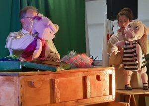 Zdjęcie wykonane podczas występu teatru. Scenografię stanowi drewniana skrzynia oraz stolik na którym dwoje aktorów opiera kierowane przez siebie lalki. Lalki imitujaa postacie dwóch świnek.