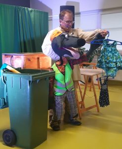 Zdjęcie wykonane podczas przedstawienia teatru. Aktor podrzymuje dużą kukiełkę psa ubranego w kraciaste spodnie i koszulę oraz odblaskową kamizelkę. W drugiej dłoni aktor trzyma zawieszone na wieszaku ubrania. Obok stoi zielony kosz na śmieci.