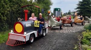 Zdjęcie kolejki z wagonikami przypominającymi dinozaury. Kolejka ciągnięta jest przez lokomotywę prowadzoną przez Pana. Obok niego siedzi jedna z nauczycielek. W wagonikach siedzą uczniowie oddzialu przedszkolnego.