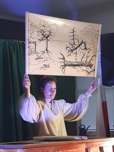Zdjęcie wykonane podczas przedstawienia teatru. Aktorka trzyma w wyciągniętych nad głową dłoniach czarno-białą grafikę przedstawioną las z przewróconymi drzewami.