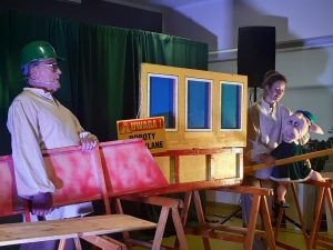 Zdjęcie wykonane podczas przedstawienia teatru. Scenografię ustawiono na drewnianych wspornikach. Na środku oparto przednią ścianę budynku na której zawieszono kartkę Uwaga ! roboty budowlane. Po lewej stronie stoi aktor z zielonym kaskiem na głowie trzymający fragment budynku a po prawej aktorka z kukiełką świnki ubraną w zieloną sukienkę i trzymającą poziomice.