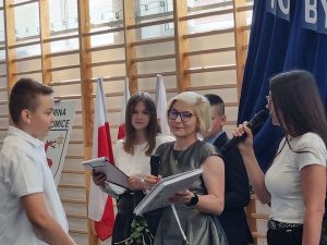 Pani Dyrektor wręcza książkę uczniowi klasy ósmej, który otrzymał specjalne wyróznieniew konkursie plastyczno-recytatorskim. Obok Pani Dyrektor stoi wychowawczyni ucznia mówiąca przez mikrofon.