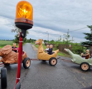 Uczniowie jeżdżą w połączonych ze sobą wagonikach na kółkach przypominających dinozaury. Na pierwszym planie widać rurkę z przyczepioną do niego lampką imitująco kogut służb ratunkowych.
