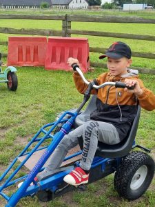 Chłopiec siedzi na małym pojeździe przypominającym rower. Uczeń trzyma ręce na kierownicy a nogi na pedałach. Zdjęcie wykonane na zielonym pastwisku.