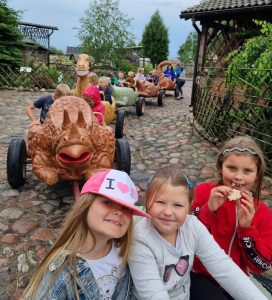 Trzy uczennice oddziału przedszkolnego pozują do zdjęcia. Jedna z nich je ciastko. W tle widać połączone wagoniki w kształcie dinozaurów, w których siedzą dzieci.