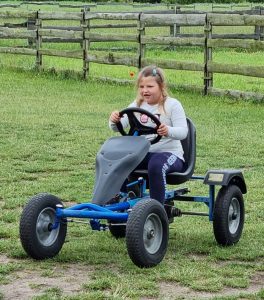 Dziewczynka siedzi na małym pojeździe przypominającym traktor. Zdjęcie wykonane na zielonym pastwisku.