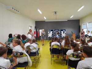 Pani Dyrektor wręcza świadectwo jednemu z uczniów, który do niej podchodzi. Obok stoi wychowawczyni klasy III z jednym ze swoich uczniów. Na zdjęciu widać głowy siedzących na krzesłach pozostałych uczniów.
