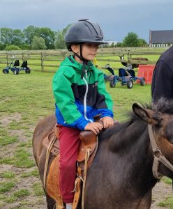 Uczeń oddziału przedszkolnego siedzi na koniu. Na głowie ma kask a rękoma trzyma się siodła. W tle widać zielone pastwisko i pojazdy do jeżdżenia dla dzieci.