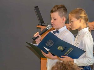 Dwoje uczniów prowadzących zakończenie roku szkolnego stoi z otwartymi teczkami z napisem absolwent szkoły podstawowej. Chłopiec trzyma w ręku mkrofon. Zdjęcie wykonane w zbliżeniu.