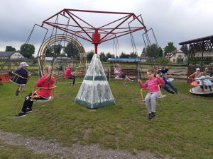 Uczniowie oddziału przedszkolnego podczas zabawy na łańcuchowej karuzeli. Dzieci trzymają się rękoma łańcuchów, do których przyczepione są krzesełka.