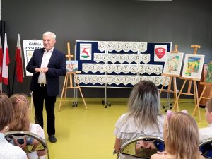 Wójt Gminy Łysomice podczas przemowyna rozdaniu nagród konkursowych. W tle stoi tablica z napisem GMINNY KONKURS PLASTYCZNO-RECYTATORSKI.