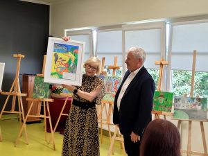 Pani Dyrektor podnosi w górę konkursową pracę ucznia, której przyznała specjalną nagrodę. Obok stoi wójt przyglądający się rysunkowi.