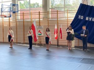 Zdjęcie nowego pocztu flagowego. Uczniowie mają przełożone biało-czerwone szarfy a uczeń stojący po środku trzyma flagę Polski. Obok stoją prowadzący galę zakończenia roku.