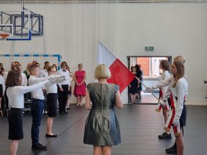 Zdjęcie wykonane podczas ślubowania nowego pocztu flagowego. Po środku odwrócona plecami stoi Pani Dyrektor. Po jej prawej stronie z opuszczoną flagową stoi odchodzący poczet flagowy a po lewej ucziowi przejmujący poczet z podniesioną w górę lewą dłonią.
